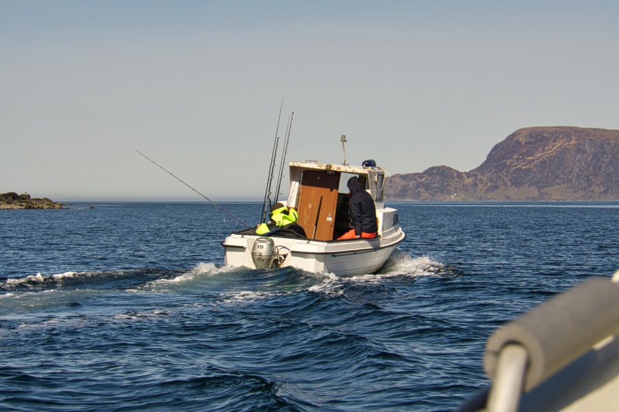 En liten fiskebåt med to personer i arbeidsklær kjører utover på åpent hav med fiskestenger om bord, med fjell og kystlandskap i bakgrunnen.