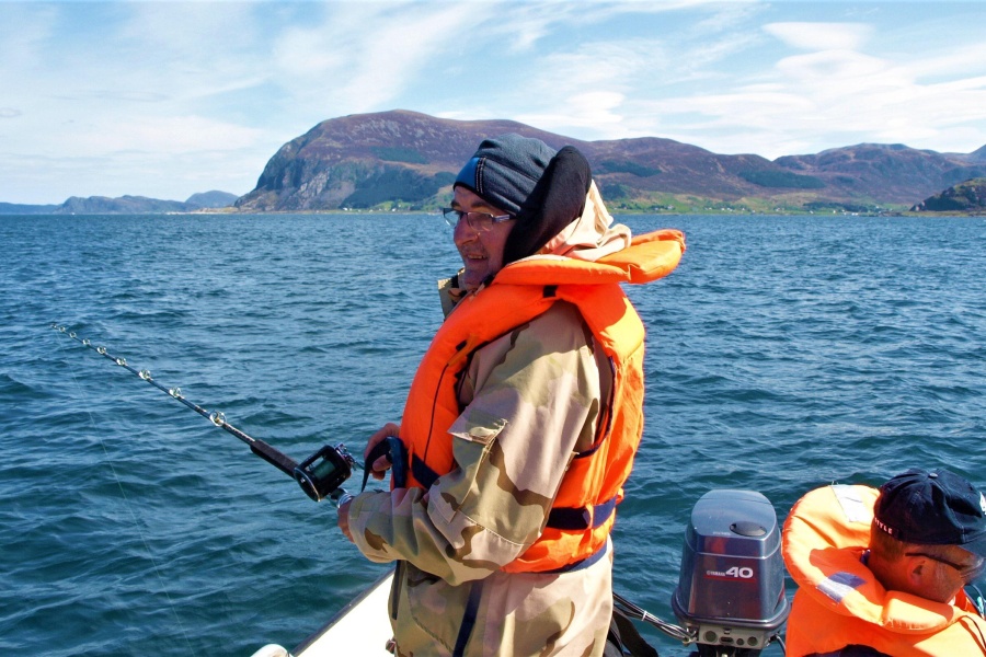 To personer fisker fra en båt på åpent hav omgitt av fjell og blå himmel, iført redningsvester.