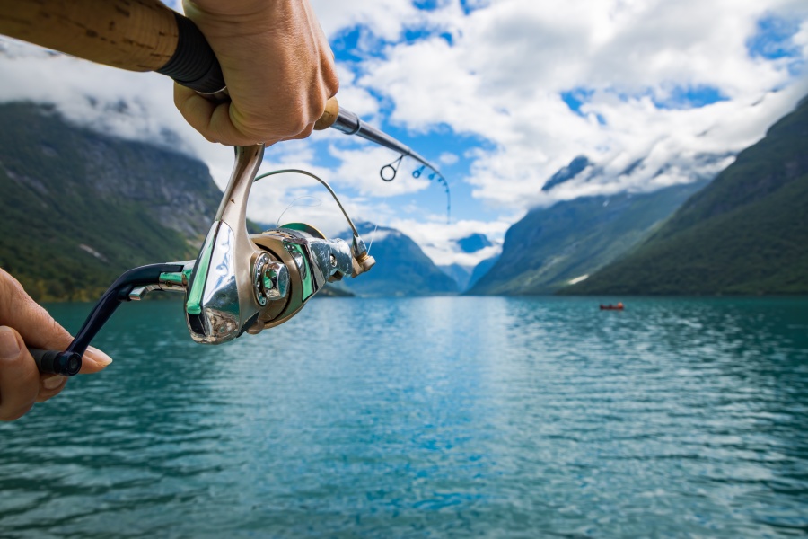 En person fisker med en snell i klart vann omgitt av fjell og solskinn, trolig i en norsk fjord.