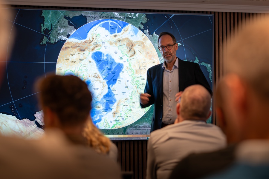 Foredragsholder peker og forklarer foran et stort kart over Arktis på en skjerm, med publikum sittende i forgrunnen.