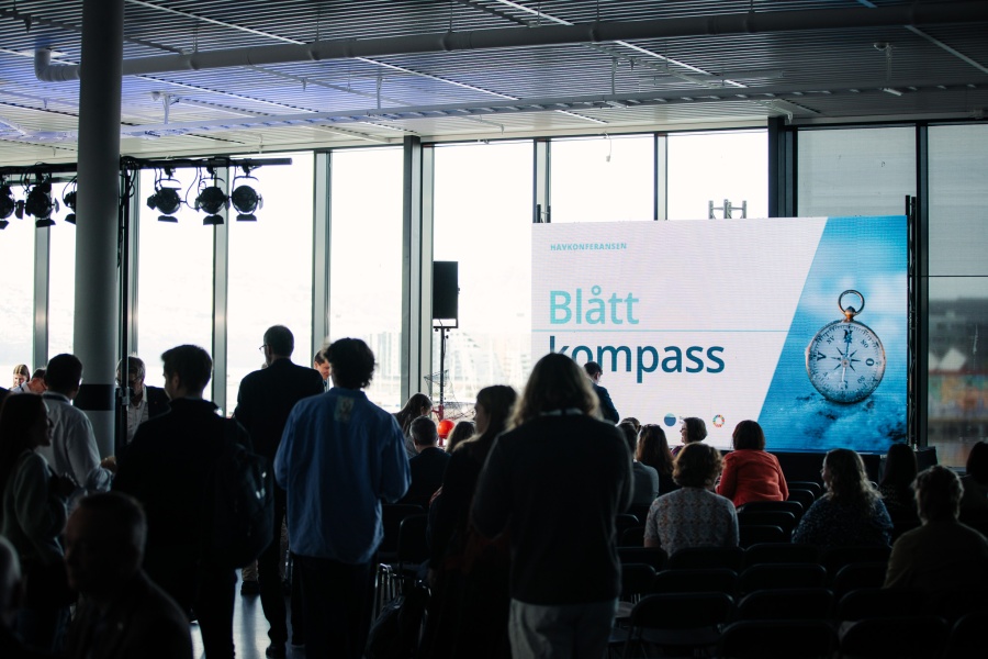 Publikum i en sal. En skjerm med et bilde som sier "Blått kompass"