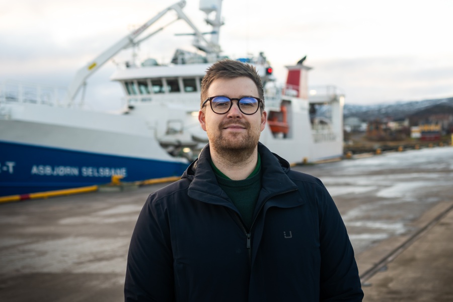 En person med kort hår og briller står utendørs på en kai. I bakgrunnen ligger et stort skip fortøyd ved havn, med kran og overbygg synlig. Personen har på seg en mørk jakke, og det er overskyet vær.