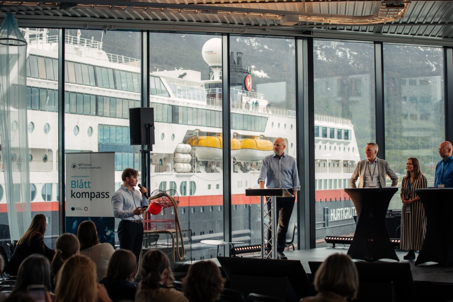 Paneldebatt på konferansen Blått kompass, med flere deltakere på scene foran store vinduer med et Hurtigruten-skip i bakgrunnen.