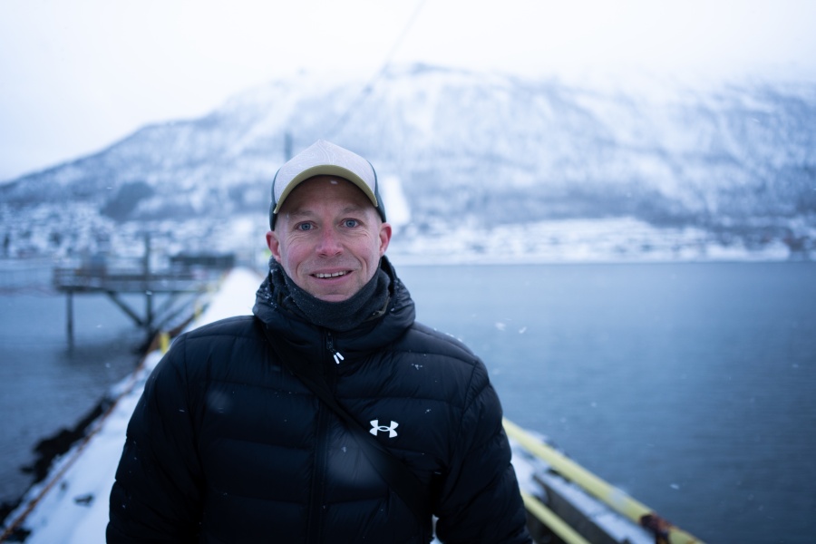 En smilende mann i vinterklær står på en brygge ved vannet, med snødekte fjell i bakgrunnen og lett snøfall rundt seg.
