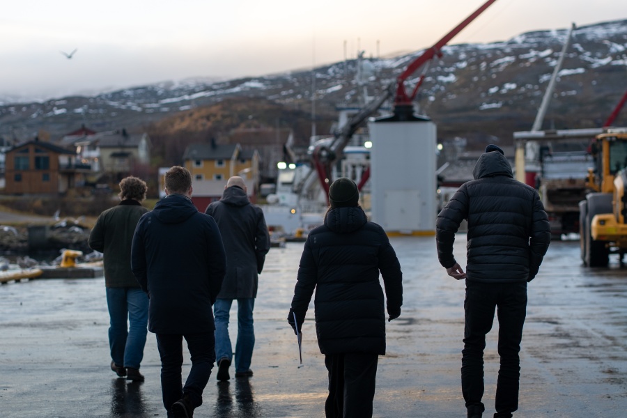 Fem personer går langs en kai i et lite kystsamfunn med snødekte fjell i bakgrunnen og industrielt utstyr rundt seg.
