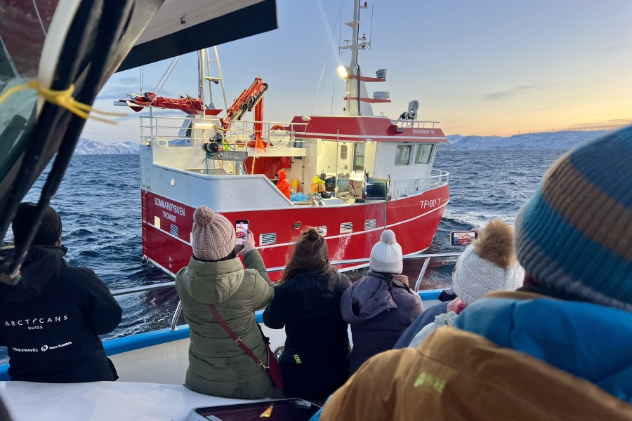 Turister i varme klær står på dekk av en båt og tar bilder av en rød fiskebåt på havet i arktisk landskap ved solnedgang.