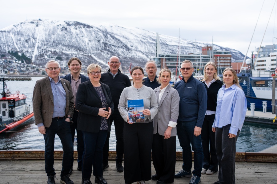En gruppe på ti personer står samlet ved en havn med båter og snødekte fjell i bakgrunnen. En kvinne i midten holder en rapport, og alle vender mot kameraet.
