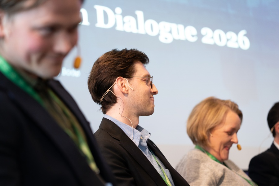 Tre personer med hodesett sitter på en scene under et arrangement, med teksten «Dialogue 2026» synlig i bakgrunnen.