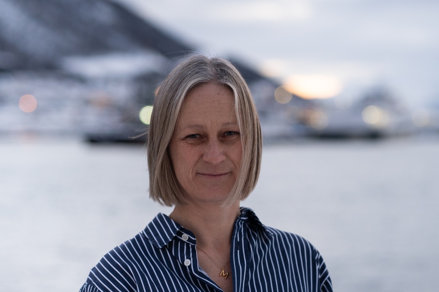 Portrett av en kvinne med kort, lyst hår som står utendørs ved vannet, med snødekte fjell og bebyggelse i bakgrunnen.