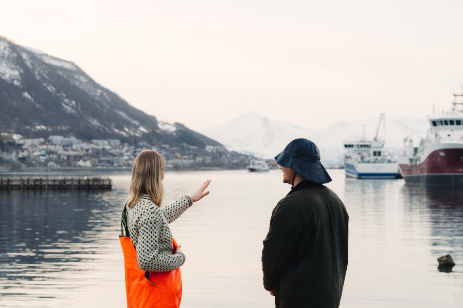 To personer står ved en havn og ser ut over vannet mot båter og snødekte fjell.