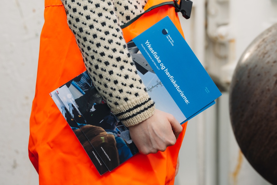 Person i oransje arbeidsklær holder en blå brosjyre med tittelen ‘Yrkesfiske og havbrukslære’, stående utendørs