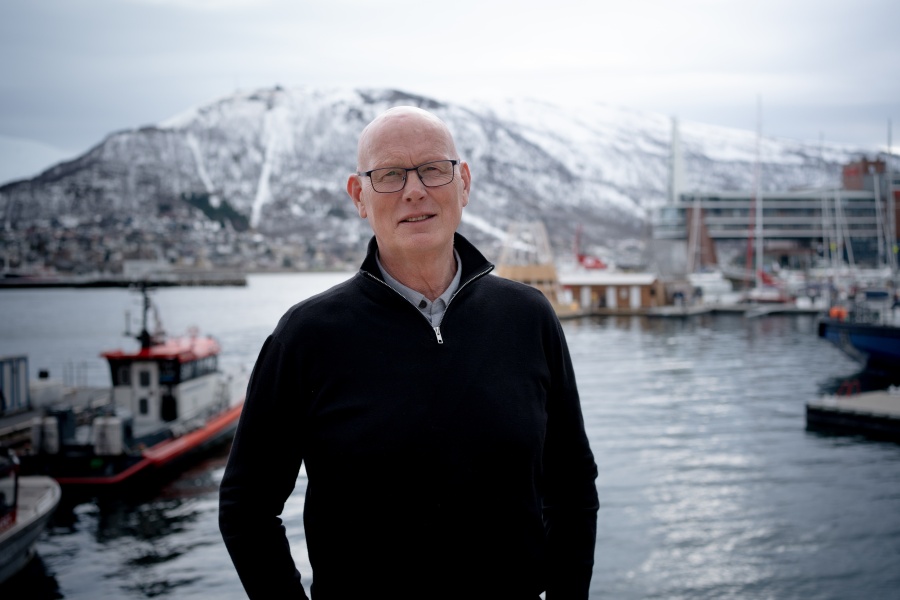 Mann med briller står ved en havn, med båter og snødekte fjell i bakgrunnen.