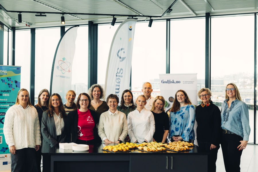 En gruppe på rundt 14 personer står samlet bak et bord med småretter i et lyst lokale, smilende mot kamera, med bannere i bakgrunnen.