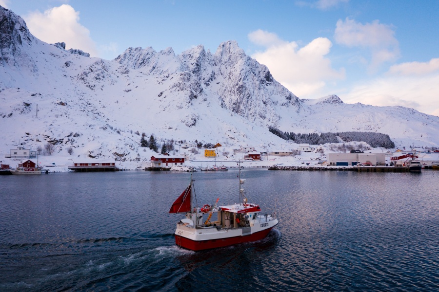 Fiskebåt på vei inn mot et snødekt kystsamfunn omgitt av bratte fjell i nordnorsk vinterlandskap.