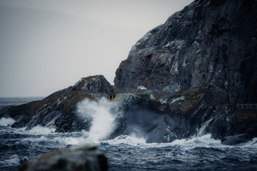 Bølger slår mot en bratt og værutsatt kystvei langs havet. En bil kjører på den smale veien mellom mørke klipper og opprørt sjø.