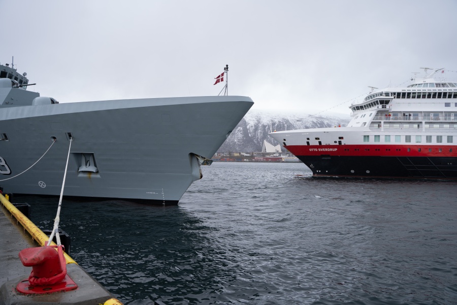 Grått marinefartøy og Hurtigruteskipet Otto Sverdrup ligger ved kai i Tromsø, med snødekte fjell i bakgrunnen.