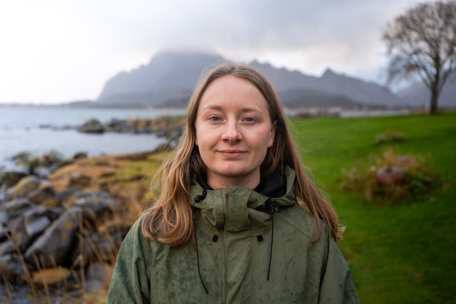 Therese Hugstmyr Woie står ved en steinete kystlinje med havet og fjell i bakgrunnen. Hun har på seg en grønn jakke og ser mot kameraet.