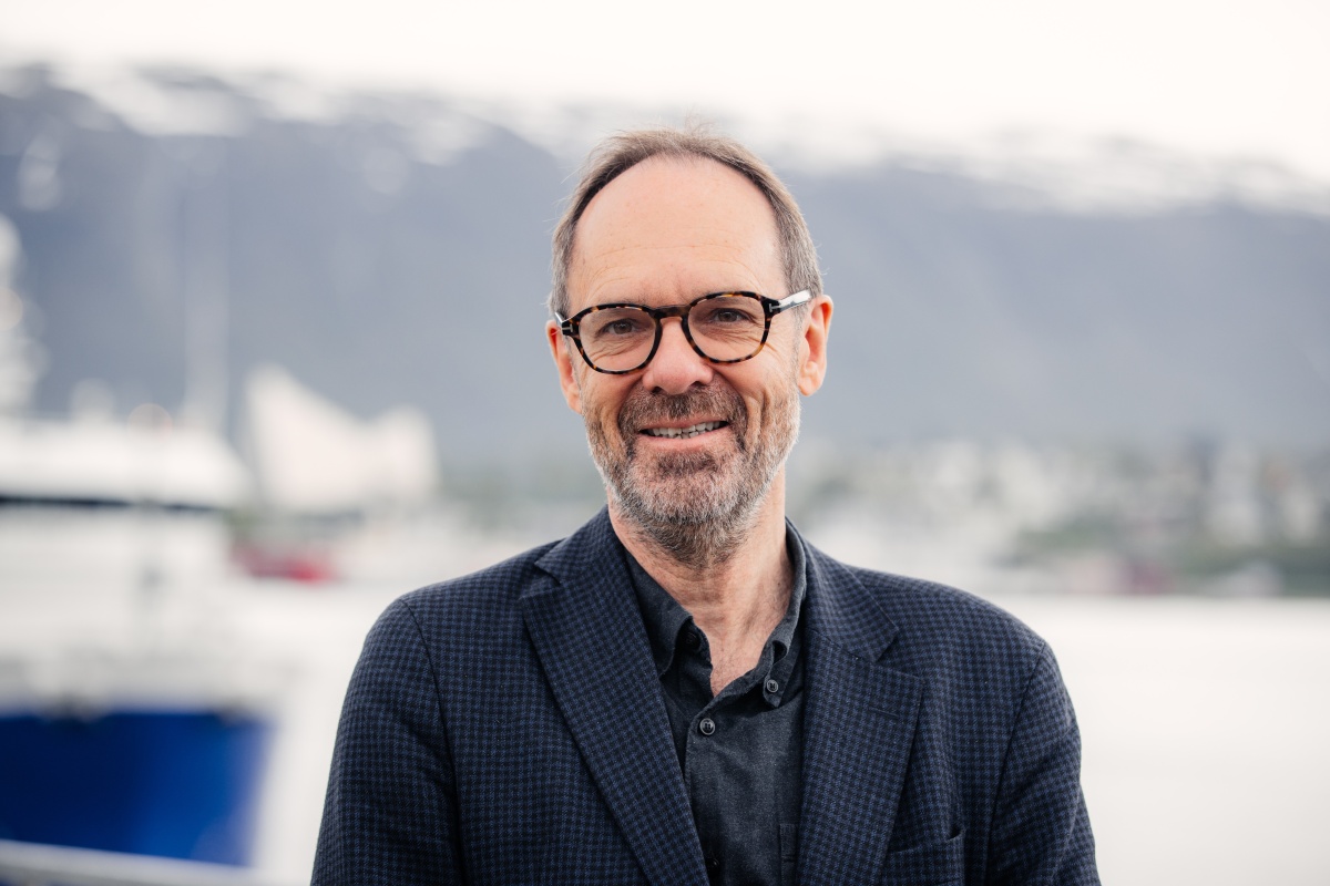 Portrett av mann i dressjakke med briller, smilende foran hav og fjell i bakgrunnen.