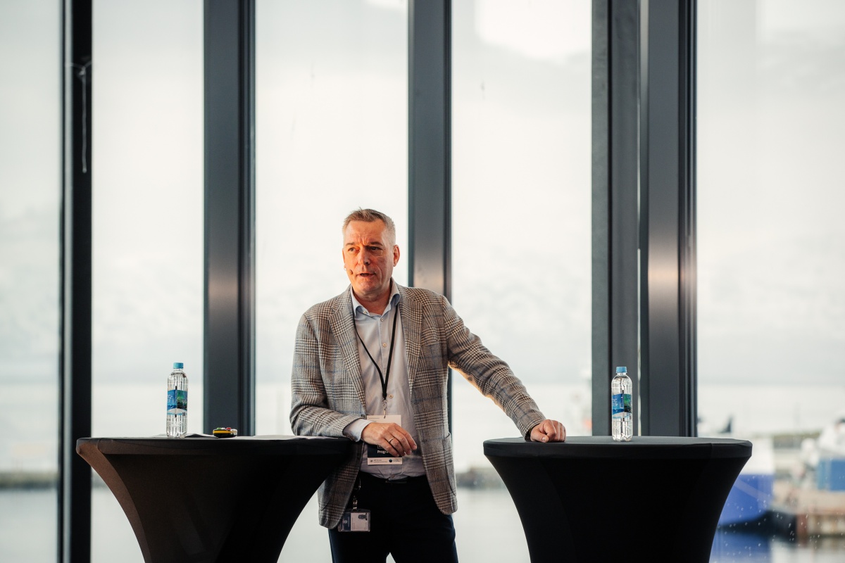 Person med mikrofon står mellom to høye bord under et arrangement, med utsikt mot hav og fjell gjennom store vinduer.