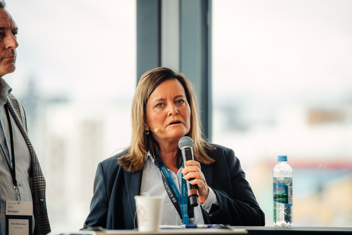 Person med mikrofon deltar i paneldebatt, iført mørk jakke og navneskilt.