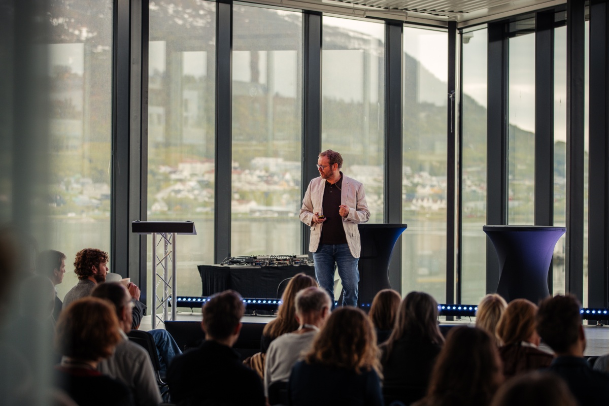 En mann holder et foredrag foran et publikum i en sal med store panoramavinduer og utsikt mot fjell og fjord. Han står på en scene med mikrofon og notater i hånden, og snakker til en lydhør forsamling. 