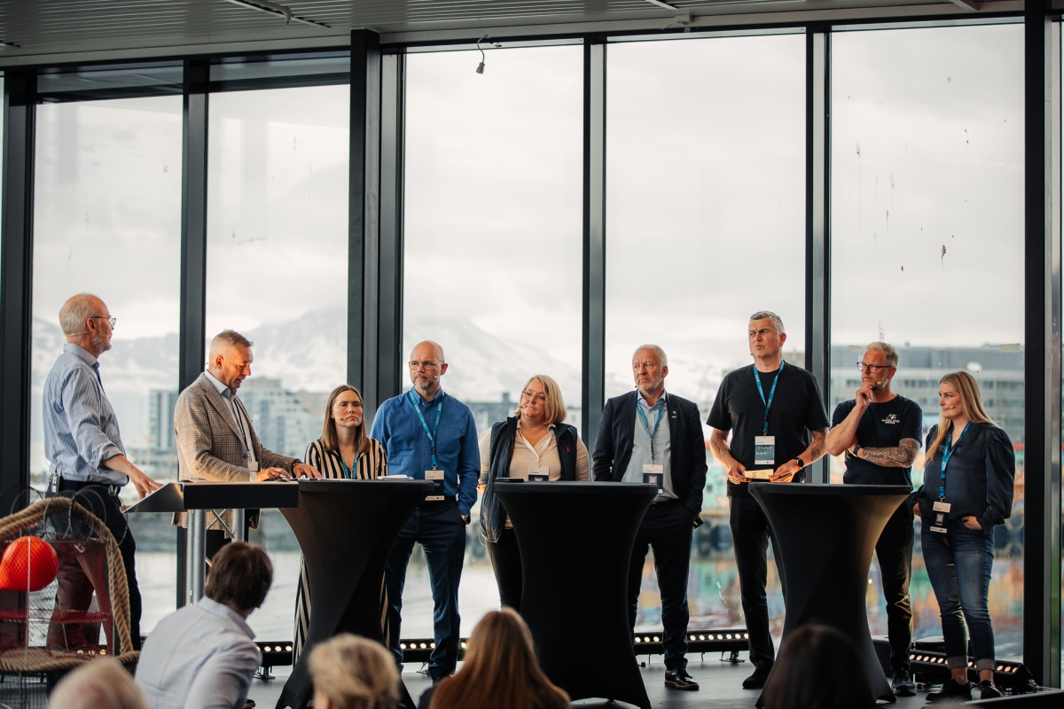 Et panel med ni personer står samlet foran store vinduer med utsikt mot fjell og by. De deltar i en panelsamtale foran publikum under en konferanse.