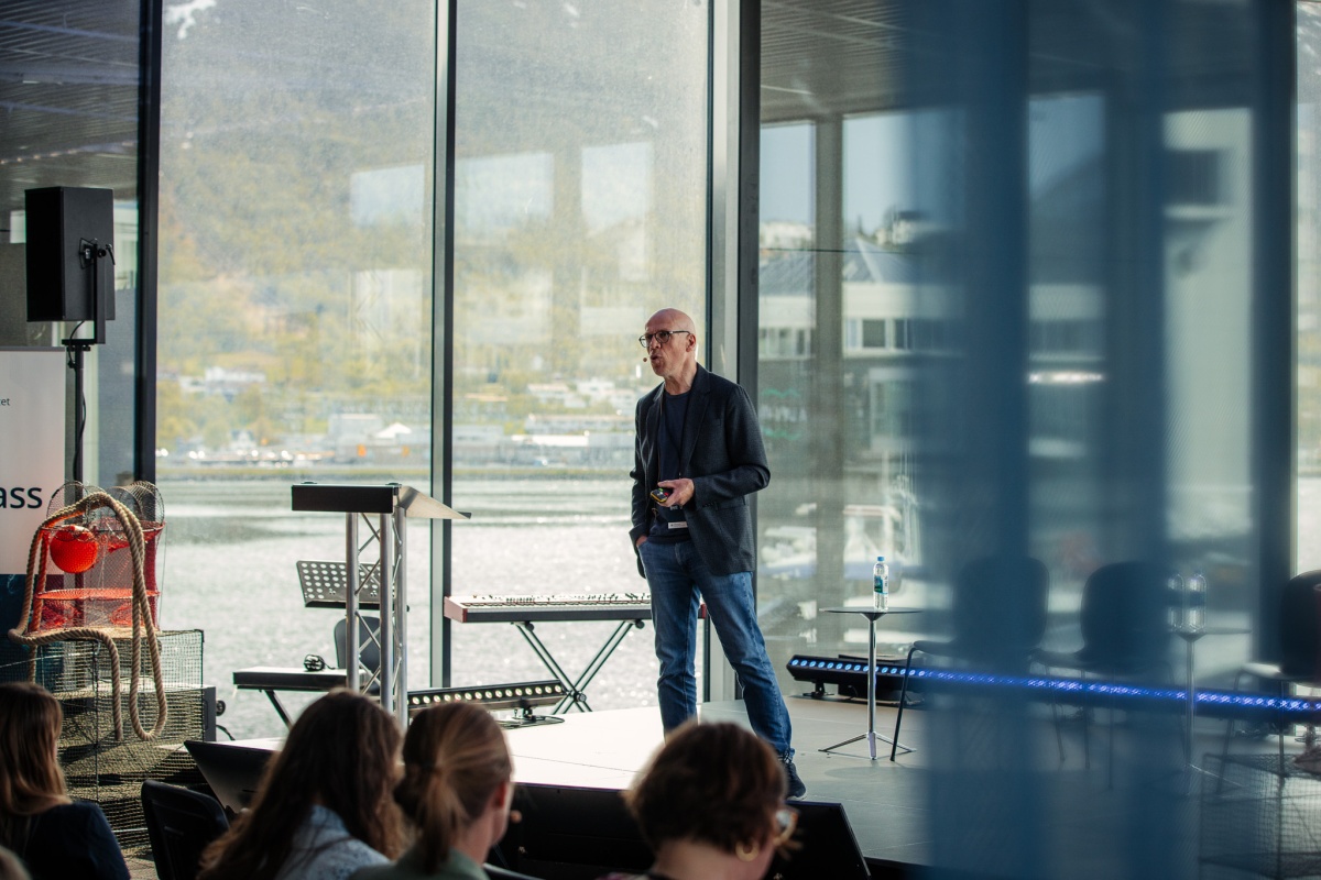 Mann med mørt blazer og skjorte står på en scene foran et publikum. Store vinduer bak han gir utsikt til sjø og fjell.