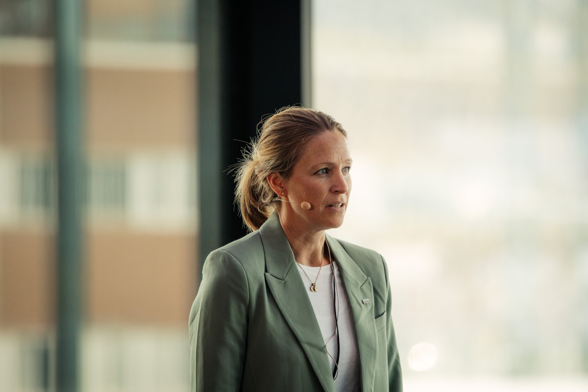 Kvinne holder innlegg iført grønn blazer og mikrofon, med store vindu og uskarp bakgrunn bak seg.