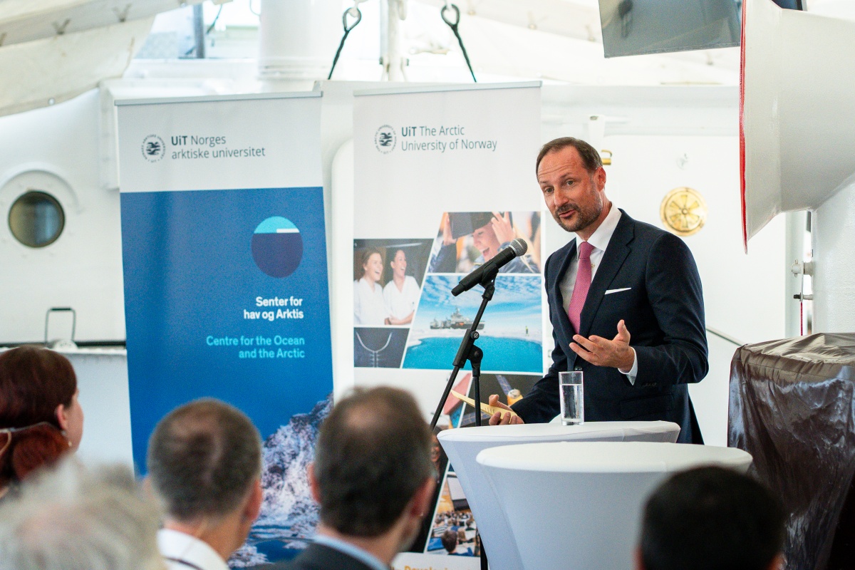 En mann i dress holder tale ved en mikrofon foran et publikum under et arrangement.