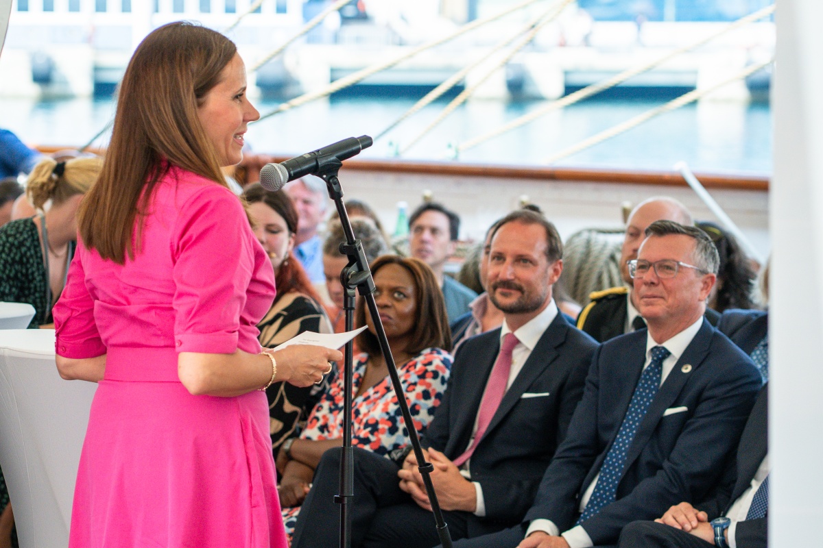 En kvinne i rosa kjole (Næss) står foran en mikrofon med ryggen til fotografen og taler til en forsamling. Rektor Olsen, Kronprins Haakon og Sanda Ojiambo i profil. 