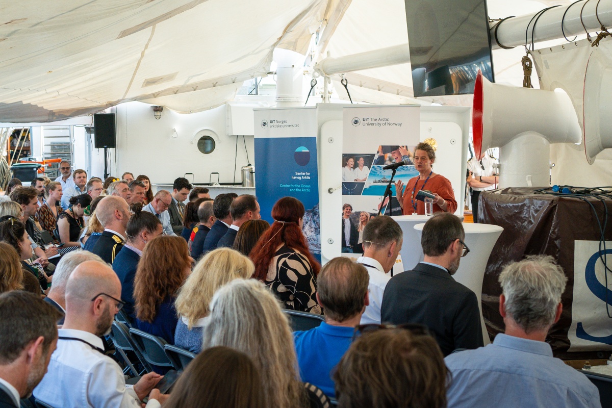 En foredragsholder står ved et podium og taler til et publikum under et arrangement arrangert av UiT Norges arktiske universitet. Publikum sitter under et telt, og bannere viser UiT-logoen og «Centre for the Ocean and the Arctic»