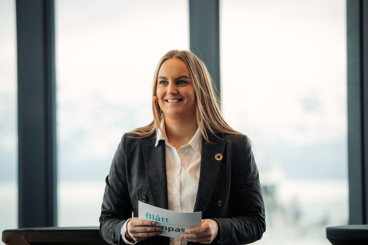 En ung kvinne med langt, lyst hår står smilende på en scene med mikrofon på kinnet. Hun har på seg hvit skjorte og mørk blazer med bærekraftsmål-pin. Hun holder et ark med teksten "Blått Kompass". I bakgrunnen skimtes store vinduer med utsikt mot fjell og hav.