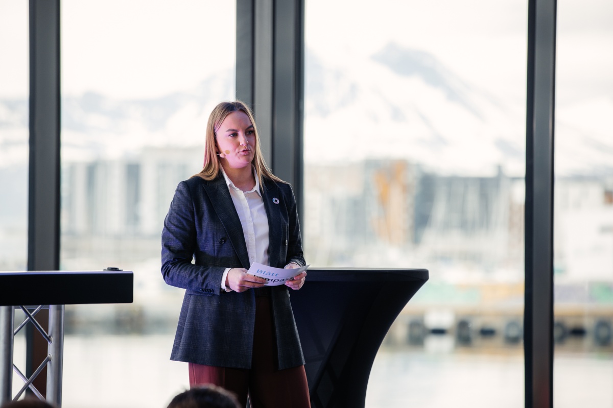 En kvinne med mikrofon står på en scene og holder et ark med teksten "Blått Kompass". Hun er kledd i blazer og hvit skjorte, og bak henne sees store vinduer med utsikt mot fjell og havn. 