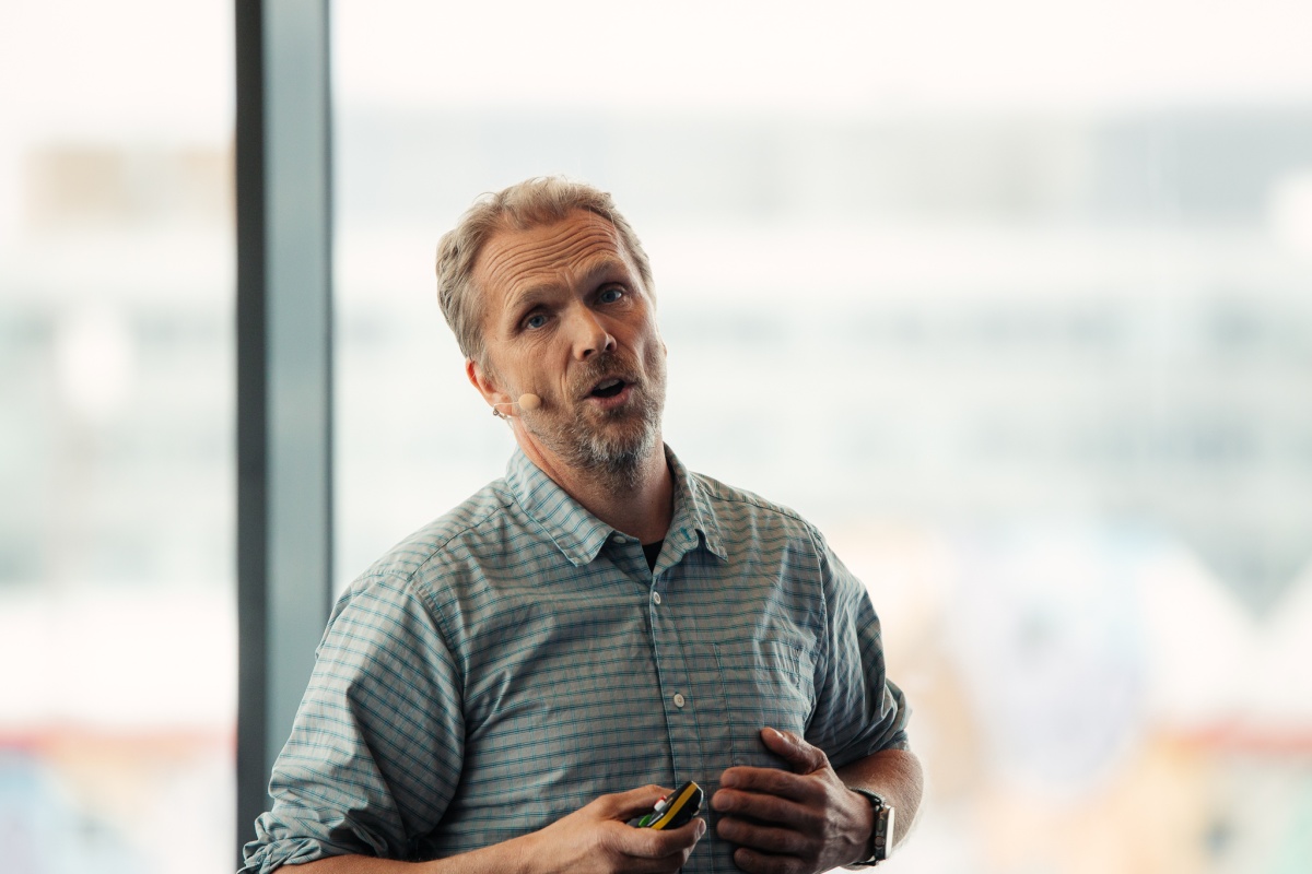 En mann holder et foredrag med mikrofon på kinnet og fjernkontroll i hånden. Han er iført en rutete skjorte og står foran et lyst bakgrunnsvindu. Han ser engasjert ut mens han snakker. 