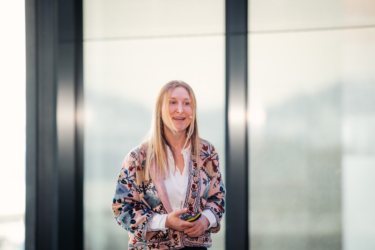 En kvinne med langt blondt hår står foran store vinduer og holder en presentasjonsfjernkontroll. Hun er iført en hvit skjorte og en mønstret jakke, og har mikrofon festet til kinnet. Hun smiler lett og ser ut til å holde et innlegg eller en presentasjon. 