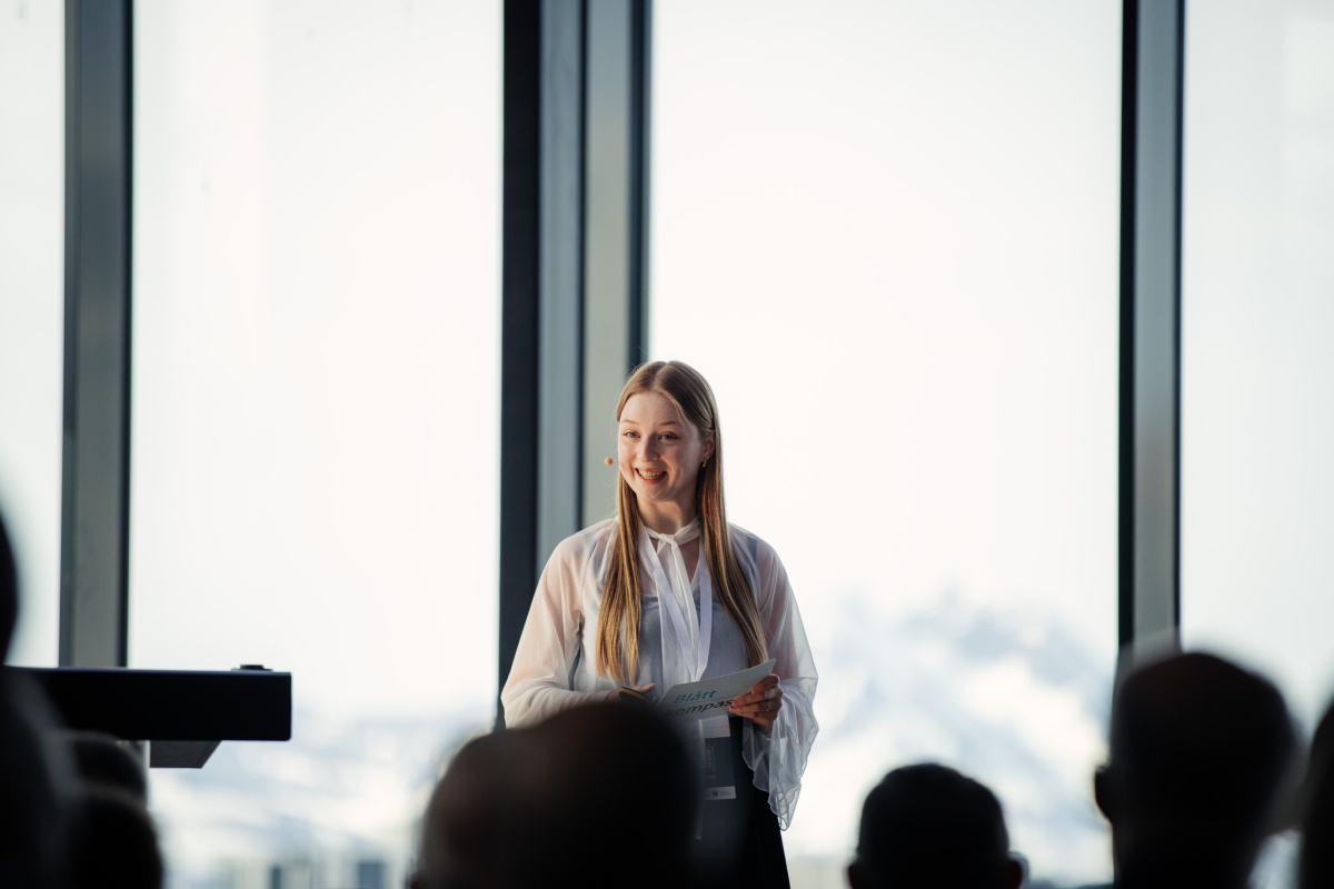 Ung kvinne står og smiler mens hun holder et notatark under et innlegg. I bakgrunnen er store vinduer og snødekte fjell