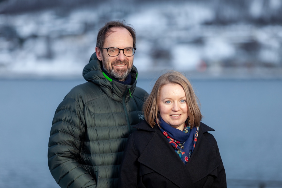 To personer står utendørs foran sjø og snødekte fjell, en mann i grønn dunjakke og briller og en kvinne i mørk kåpe og skjerf.