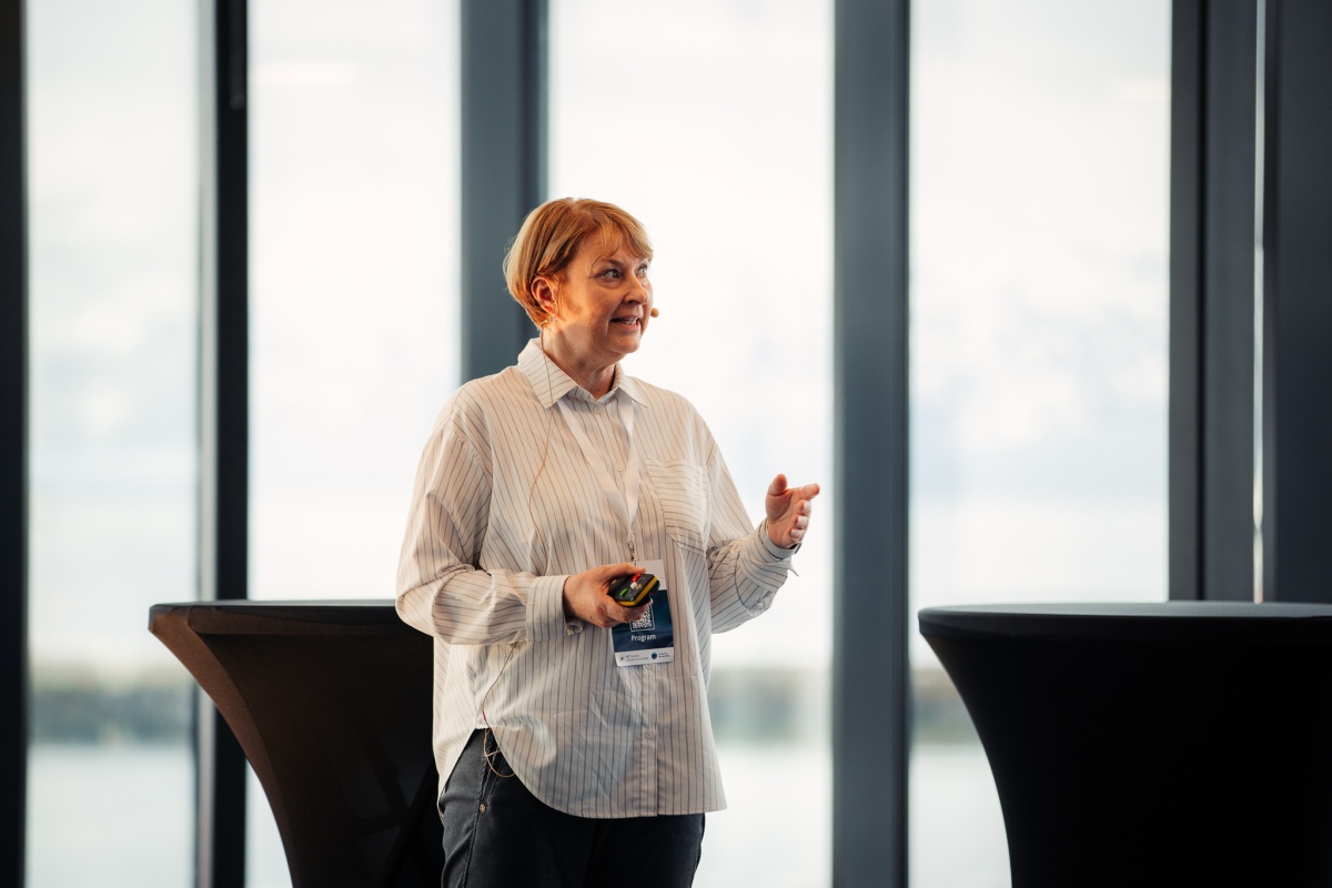 En kvinne med mikrofon holder en presentasjon med store vinduer og utsikt mot hav og fjell i bakgrunnen. 