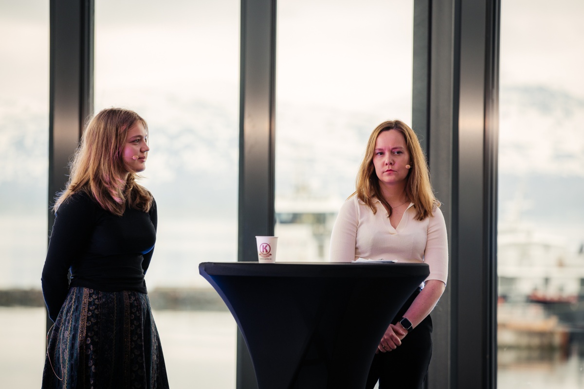 To unge kvinner står på en scene bak et høyt rundt bord under en paneldiskusjon. Begge har mikrofoner og ser konsentrerte ut. I bakgrunnen sees store vinduer med utsikt over snødekte fjell og fjord. 