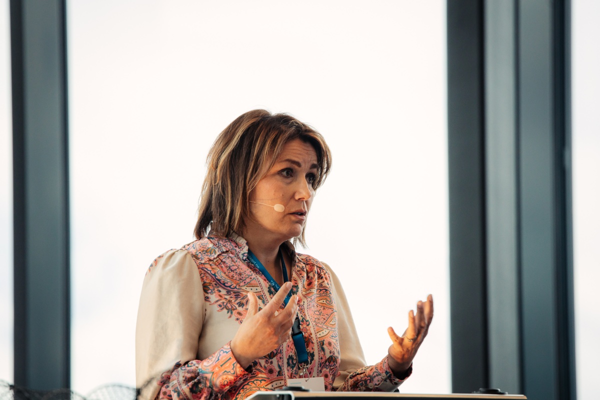 En kvinne med mikrofon står foran et stort vindu og bruker håndbevegelser for å understreke et poeng.