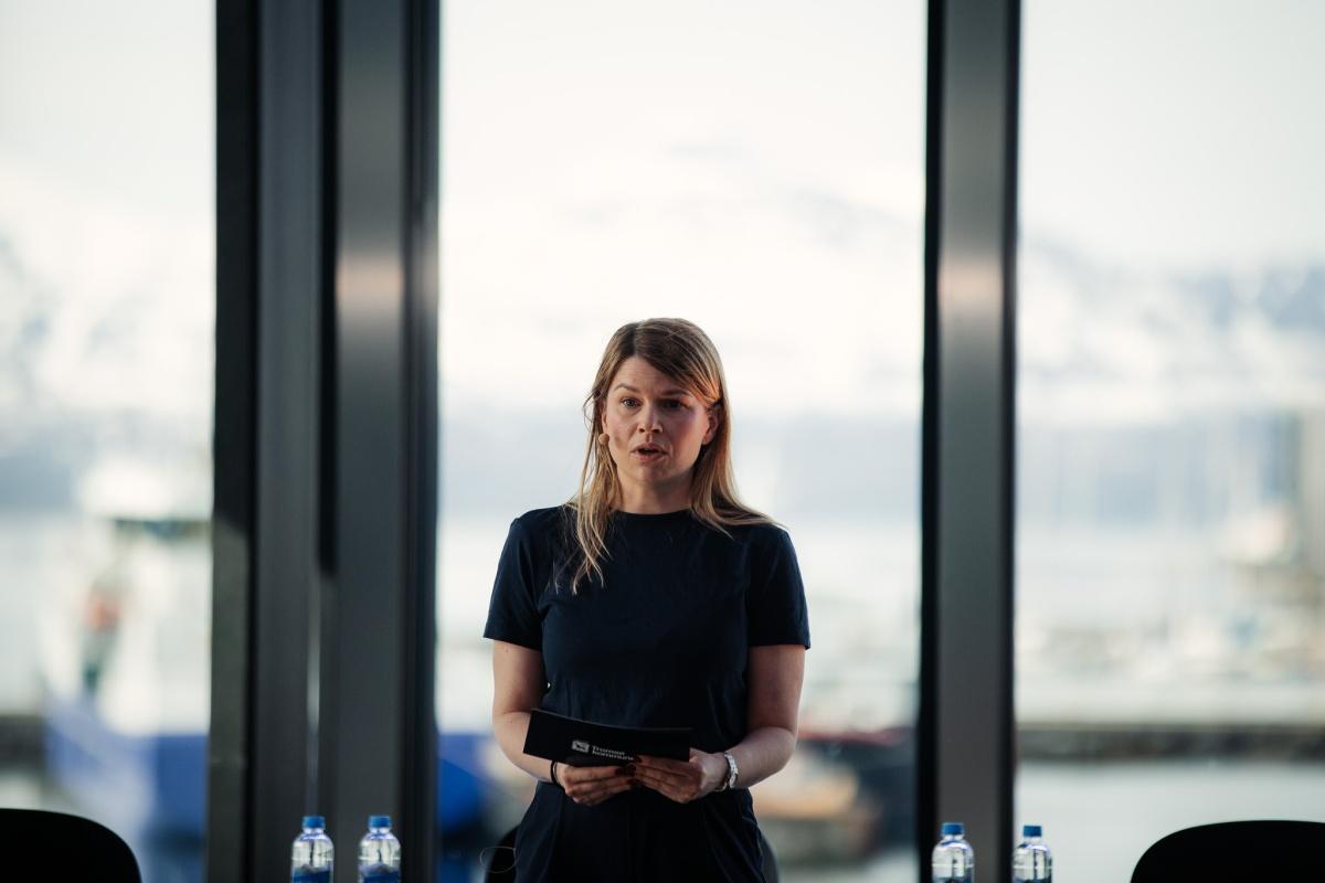Kvinne står og snakker foran store vinduer med utsikt mot havn og snødekte fjell i bakgrunnen. Hun holder et kort og er midt i et innlegg.