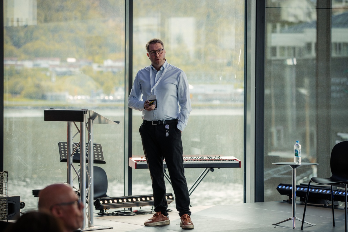 Mann med briller står på scenen med mikrofon, med store vinduer og fjordlandskap i bakgrunnen.
