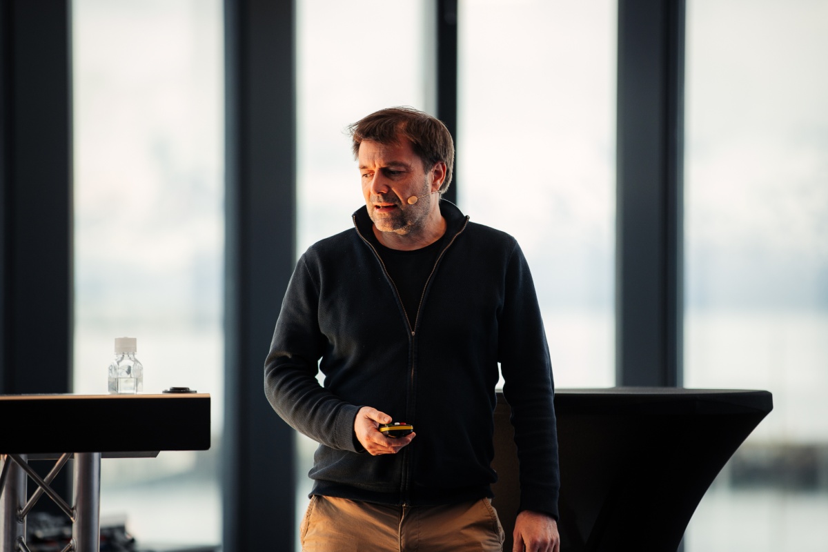 En mann med mikrofon holder et foredrag foran store vinduer med utsikt mot fjell og hav. Han står med en fjernkontroll i hånden og har fokusert blikk.