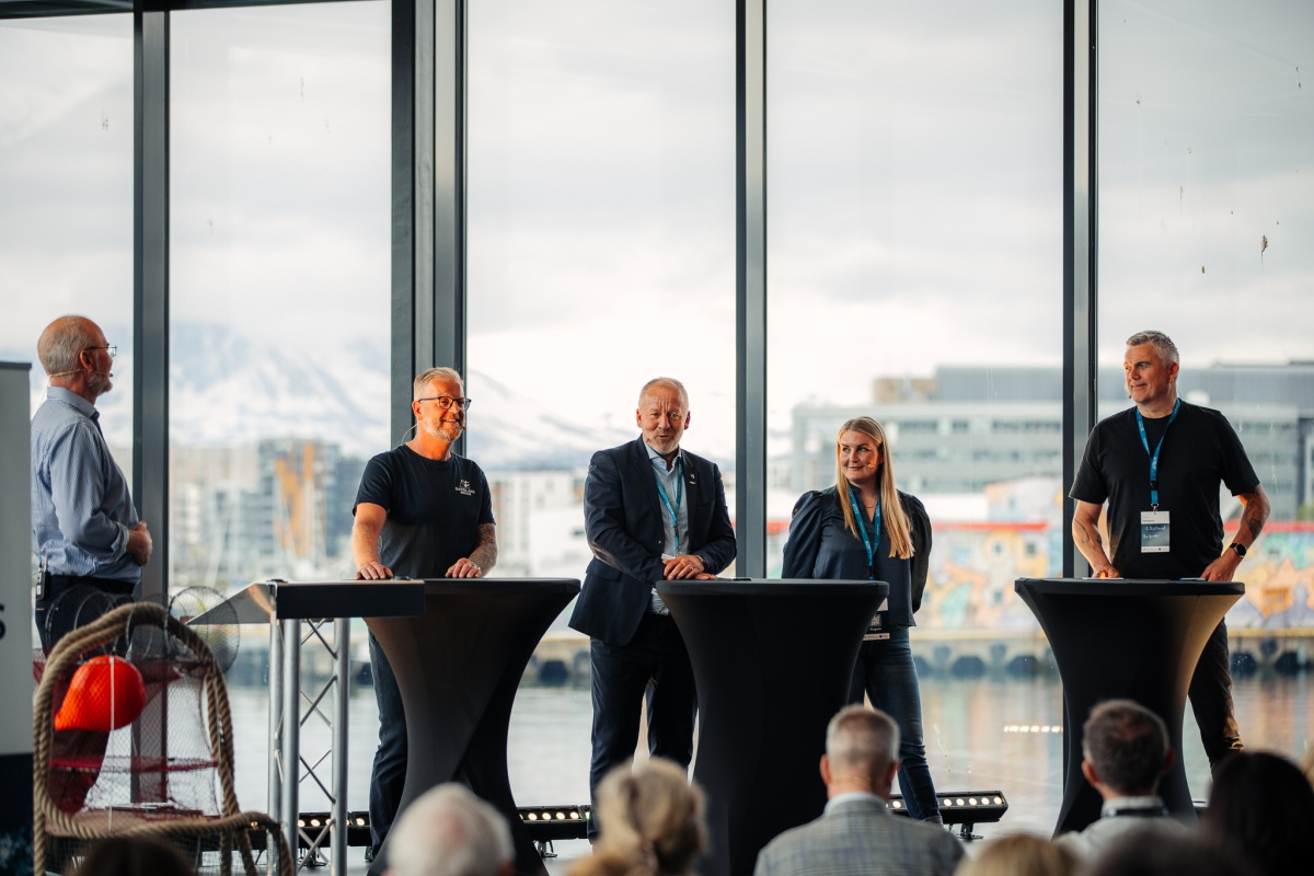 Et panel på fem personer står bak høye svarte bord og diskuterer foran et publikum. Store vinduer i bakgrunnen viser utsikt over havn og fjell. Personene har mikrofoner og én av dem snakker mens de andre lytter. Stemningen virker engasjert og profesjonell. 