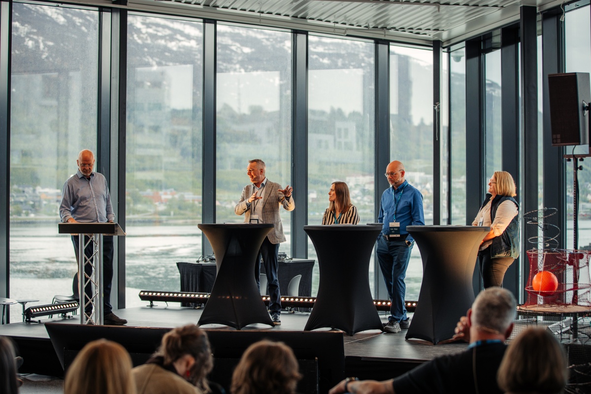 Et panel med fem personer står på en scene og deltar i en diskusjon foran publikum. De er plassert ved høye ståbord, med store vinduer bak seg som gir utsikt mot fjord og fjellandskap. Én av deltakerne gestikulerer mens han snakker, mens en annen står ved en talerstol og leser. 