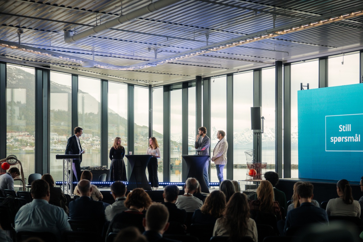 Fem paneldeltakere står på en scene foran et stort publikum, med fjell og sjø i bakgrunnen. En stor skjerm viser teksten «Still spørsmål». Paneldebatten foregår i et lyst konferanserom med panoramautsikt. 