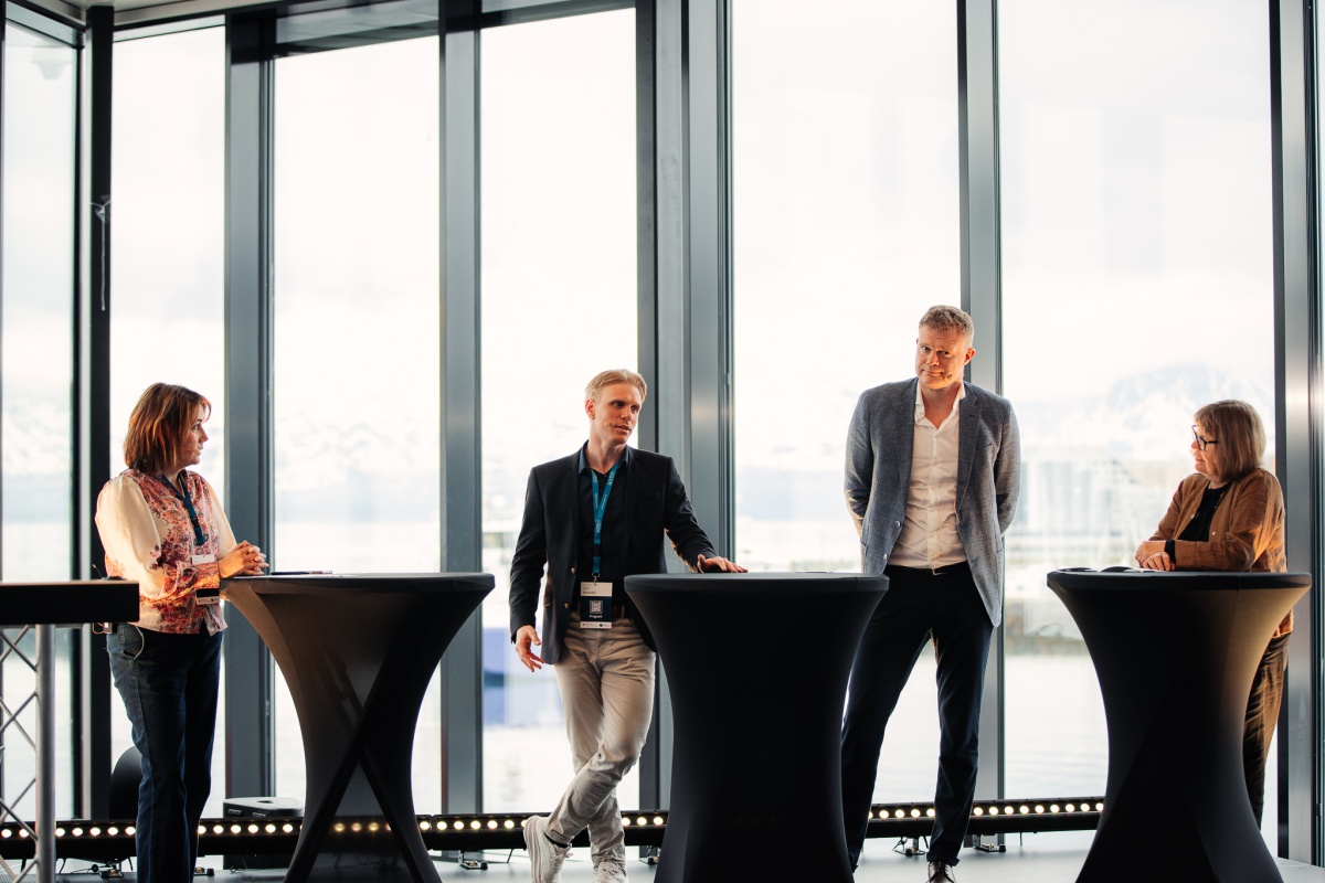 Panelsamtale mellom fire mennesker som står ved høye bord foran store vinduer med utsikt mot snødekte fjell og hav.