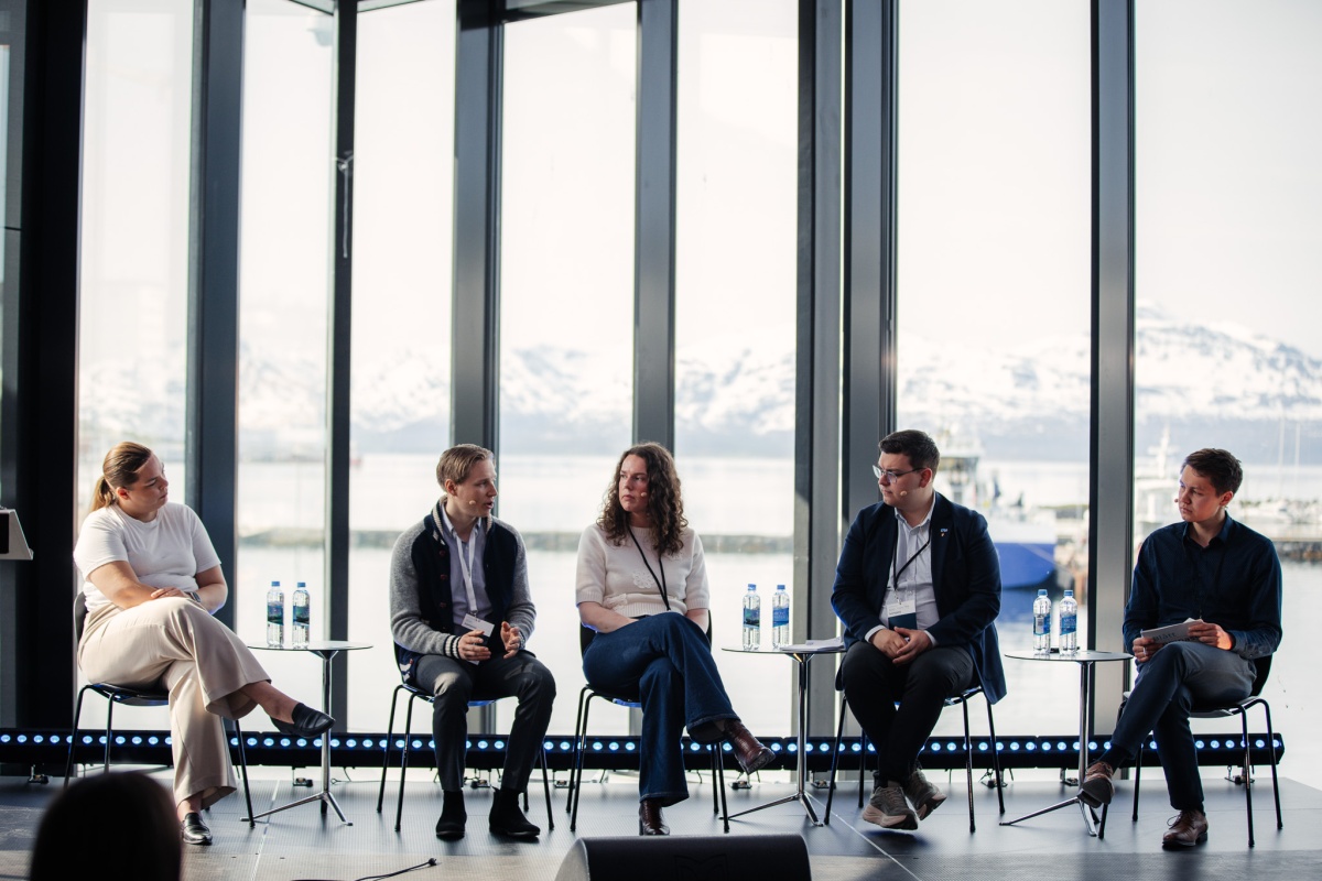 Fem personer sitter i paneldebatt foran store vinduer med utsikt til snødekte fjell. 