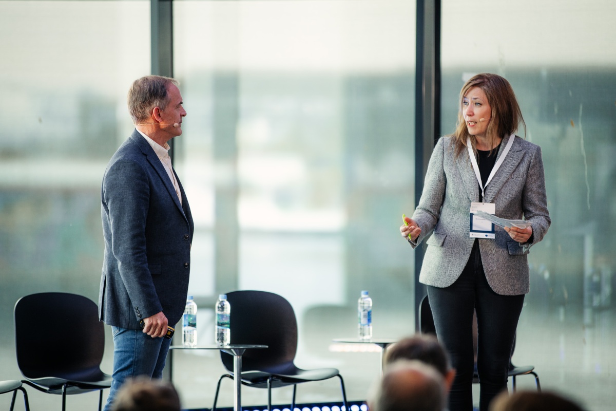 To personer står på en scene og samtaler foran publikum. Mannen til venstre holder en presentasjonsklikker, kvinnen til høyre holder et ark og snakker.