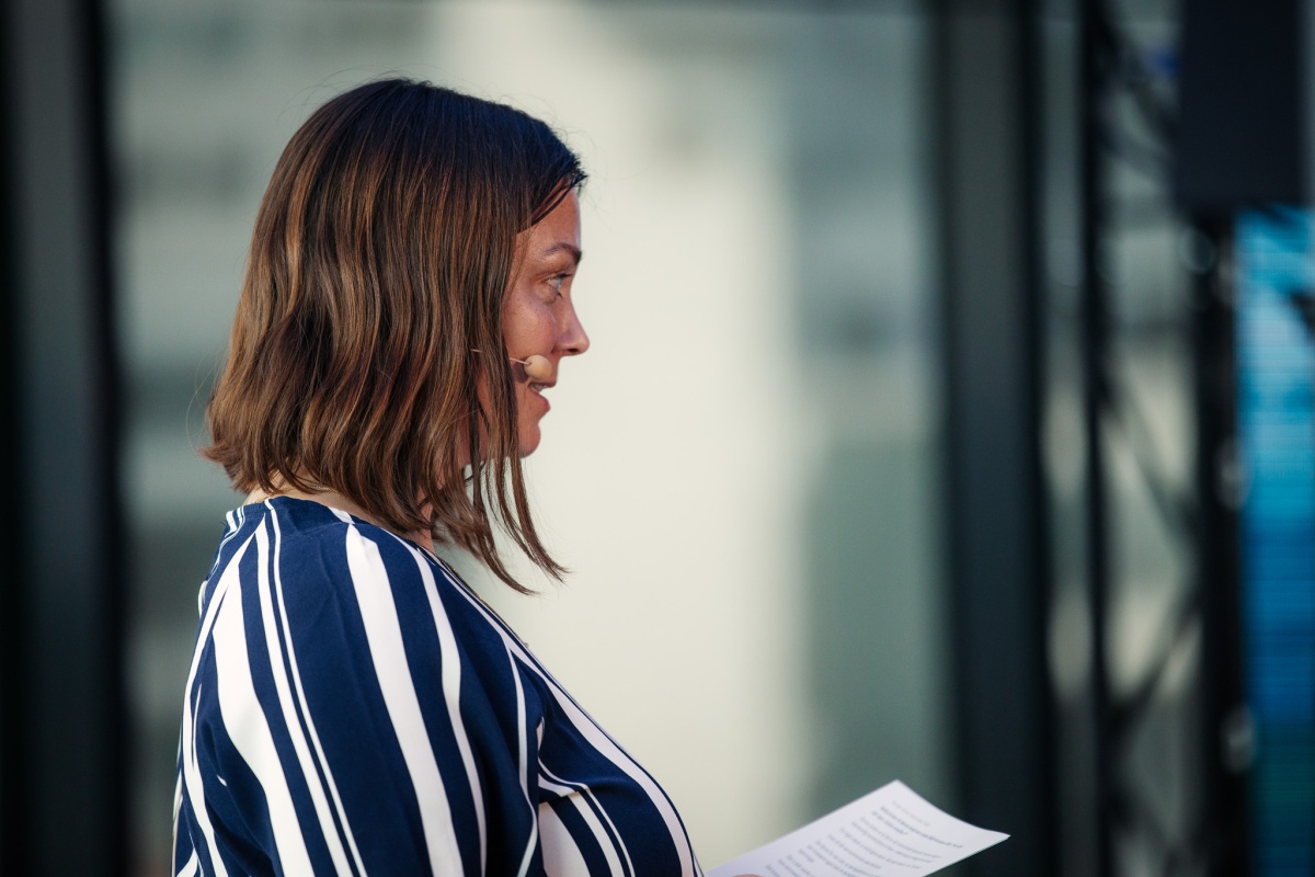 Kvinne med skulderkort hår, iført stripete bluse taler til publikum med mikrofon i ansiktet og ark i hånden